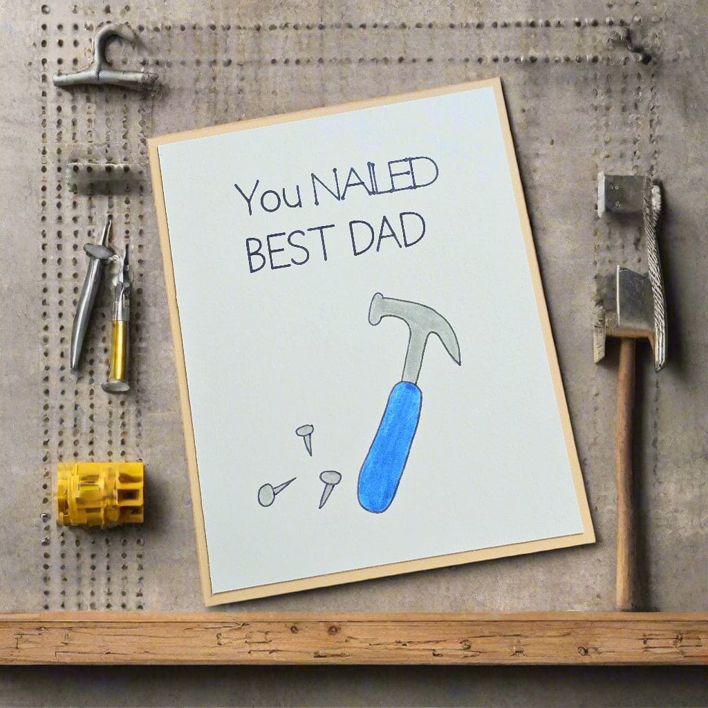 Greeting card with a blue hammer and text 'You NAILED BEST DAD' on a wooden surface.