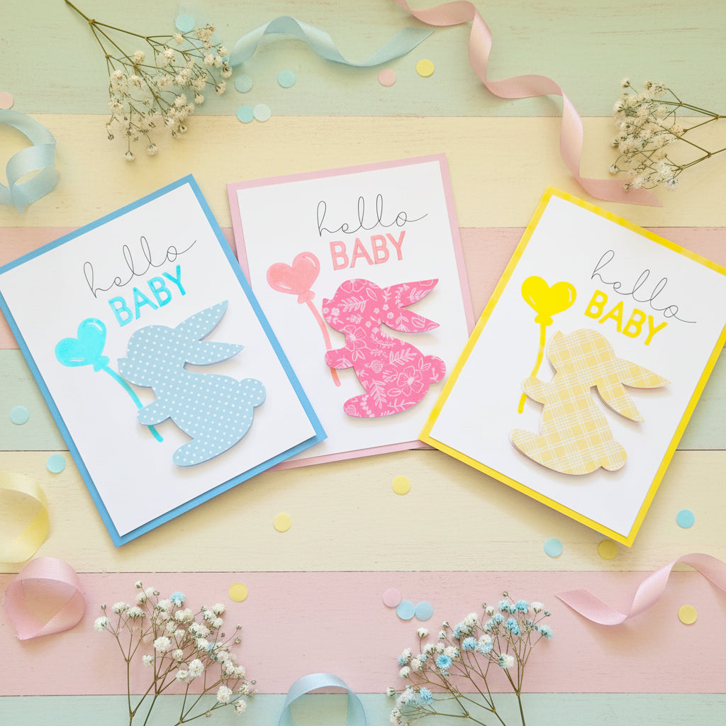 Three handmade 'hello BABY' greeting cards arranged on a light cream background. Each card features a white base with a colored border, a script 'hello BABY' text, and a dimensional bunny silhouette holding a heart-shaped balloon in different colors and patterns: light blue with white polka dots, pink with white floral print, and yellow with a white gingham pattern.