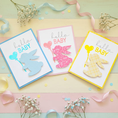 Three handmade 'hello BABY' greeting cards arranged on a light cream background. Each card features a white base with a colored border, a script 'hello BABY' text, and a dimensional bunny silhouette holding a heart-shaped balloon in different colors and patterns: light blue with white polka dots, pink with white floral print, and yellow with a white gingham pattern.