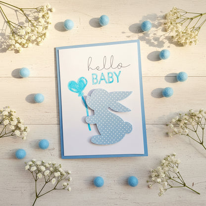 A handmade baby greeting card with a blue border, featuring the words 'hello BABY' and a layered bunny silhouette with white polka dots holding a blue heart-shaped balloon.