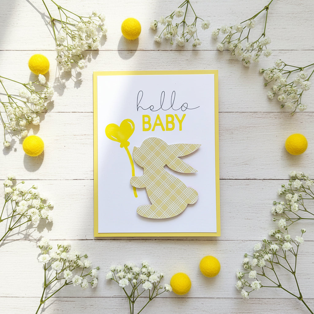 A handmade baby greeting card with a white background, yellow border, a yellow heart-shaped balloon, and a layered bunny cutout with a yellow plaid pattern. The card is displayed on a light off-white surface.