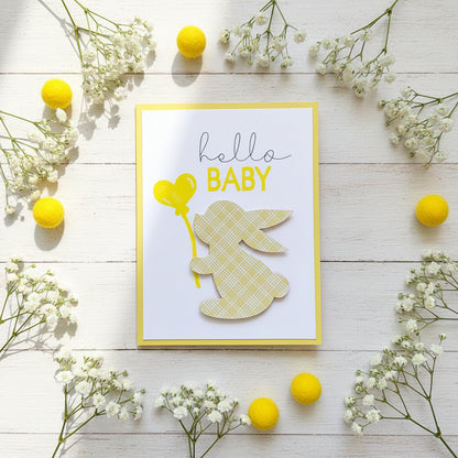 A handmade baby greeting card with a white background, yellow border, a yellow heart-shaped balloon, and a layered bunny cutout with a yellow plaid pattern. The card is displayed on a light off-white surface.