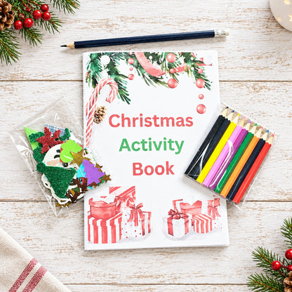 A Christmas-themed activity book titled 'Christmas Activity Book' lies on a whitewashed wooden surface, surrounded by festive props including a pencil, colored pencils, craft stickers, and evergreen branches with red berries. The book cover features holiday illustrations such as candy canes, ornaments, presents, and pine garlands.