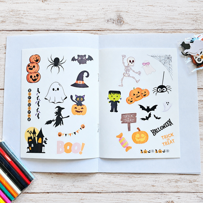 An open sticker book or sticker sheet set displayed on a light wood surface, featuring Halloween-themed stickers including pumpkins, ghosts, bats, a witch hat, Frankenstein, a haunted house, and 'Halloween' and 'Trick or Treat' text. Color pencils are visible at the bottom left, and a small packaged item is at the top right.