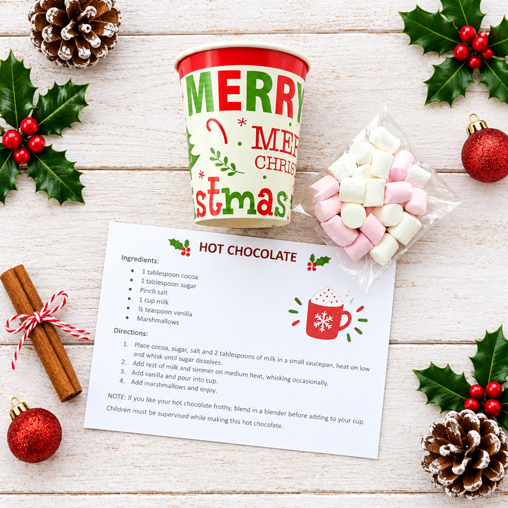 A holiday hot chocolate kit arranged on a round wooden tray over a white wood background. The set includes a red and green 'Merry Christmas' cup, a small clear bag of pink and white marshmallows, a printed recipe card titled 'HOT CHOCOLATE,' and a metal mug.