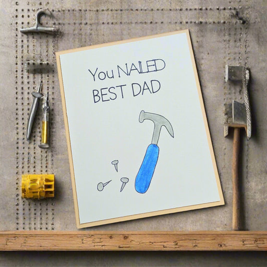 Greeting card with a blue hammer and text 'You NAILED BEST DAD' on a wooden surface.