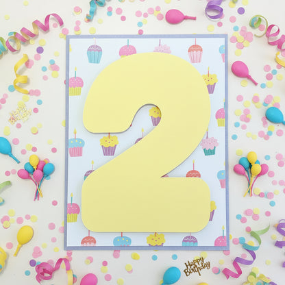 A square birthday party table number card featuring a large yellow paper '2' on a white background with a colorful cupcake pattern, displayed on a wooden table.