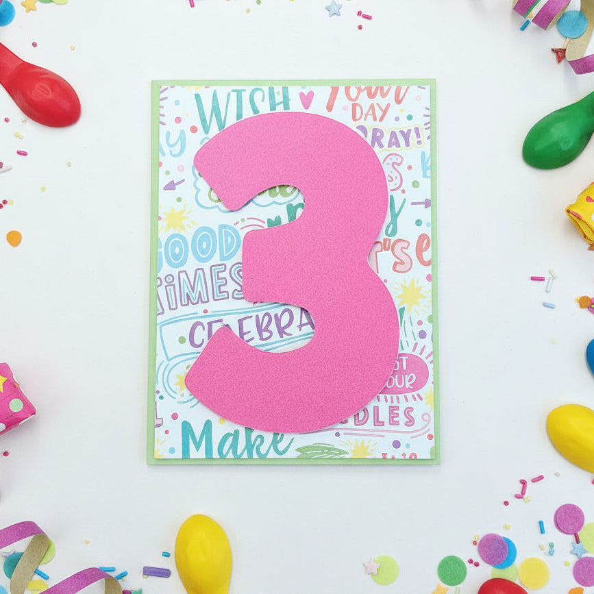 A handmade birthday card featuring a large, hot pink paper number '3' layered over a colorful retro-themed card with pastel text and doodles, displayed on a wooden tabletop.