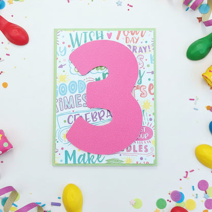 A handmade birthday card featuring a large, hot pink paper number '3' layered over a colorful retro-themed card with pastel text and doodles, displayed on a wooden tabletop.