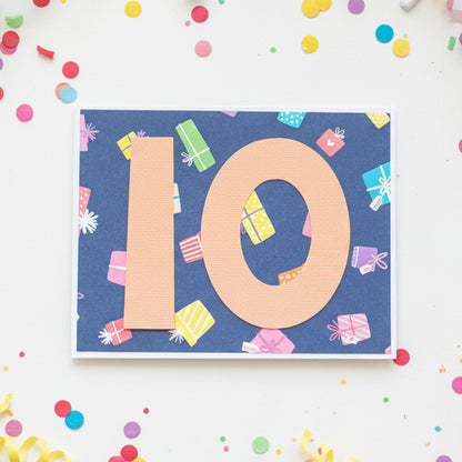 A square greeting card with a navy blue background, featuring the number 10 in large peach letters and decorated with small colorful gift boxes, placed on a wooden surface.