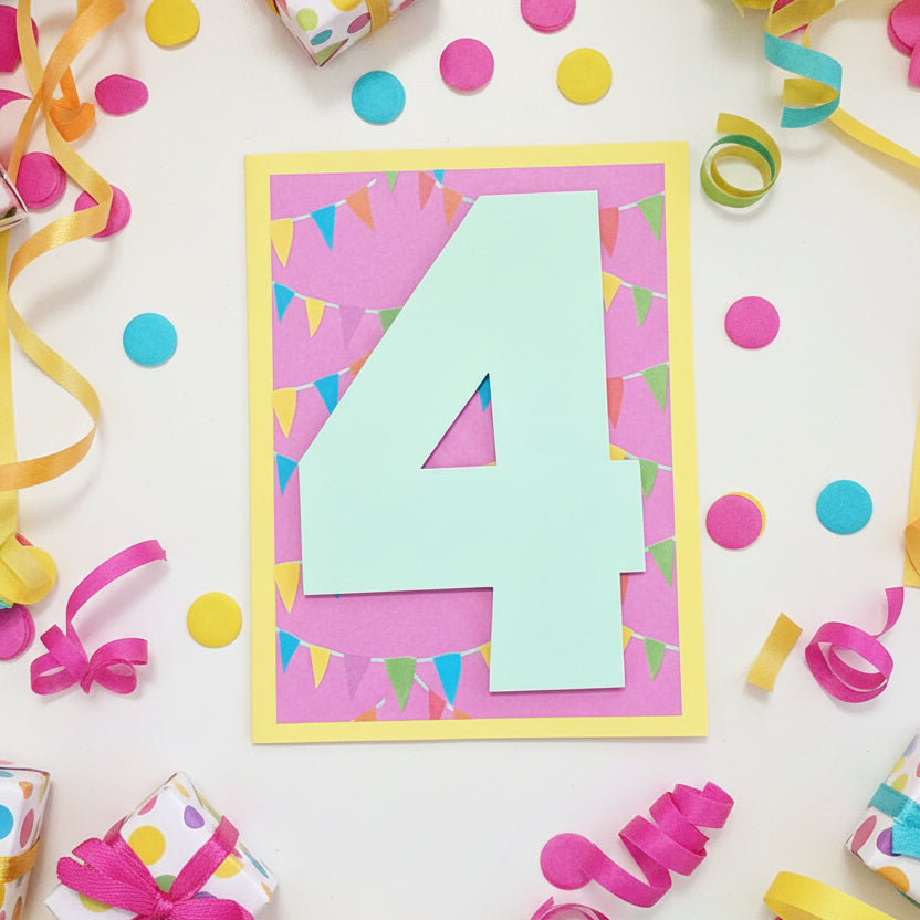 A handmade layered paper card featuring a large light blue number 4 on a pink bunting-patterned background, bordered by a yellow frame, displayed on a wooden surface.