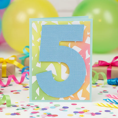 A handmade layered paper card featuring a large textured light blue number 5 on a colorful, patterned background, placed on a wooden surface.