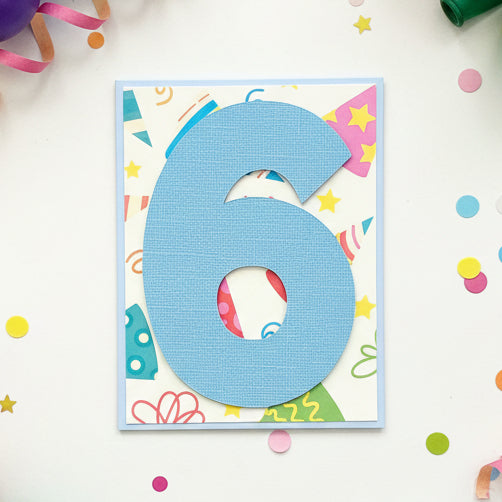 A handmade birthday card featuring a large textured light blue number 6 layered over a colorful, patterned background with party hats, stars, and doodles, displayed on a wooden surface.