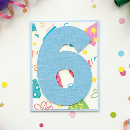 A handmade birthday card featuring a large textured light blue number 6 layered over a colorful, patterned background with party hats, stars, and doodles, displayed on a wooden surface.
