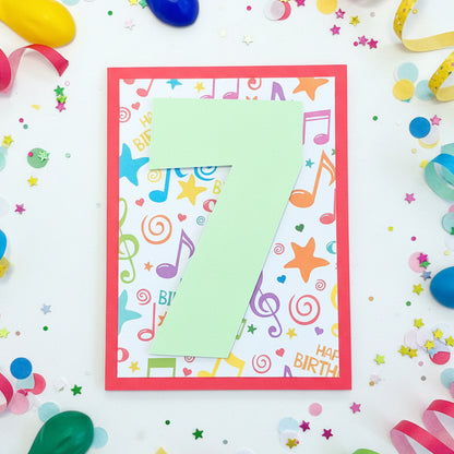 A handmade square birthday card with a large light green paper number 7 layered over a colorful music-themed background, placed on a wooden surface.