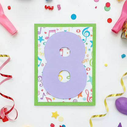 A handmade paper birthday card featuring a large lavender number 8 on a white background with colorful musical notes and stars, bordered by a green frame, placed on a wooden surface.