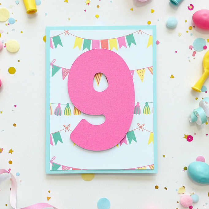 A handmade birthday card featuring a large pink foam number '9' layered over a white card with a light blue border and colorful bunting garland illustrations, displayed on a wooden tabletop.