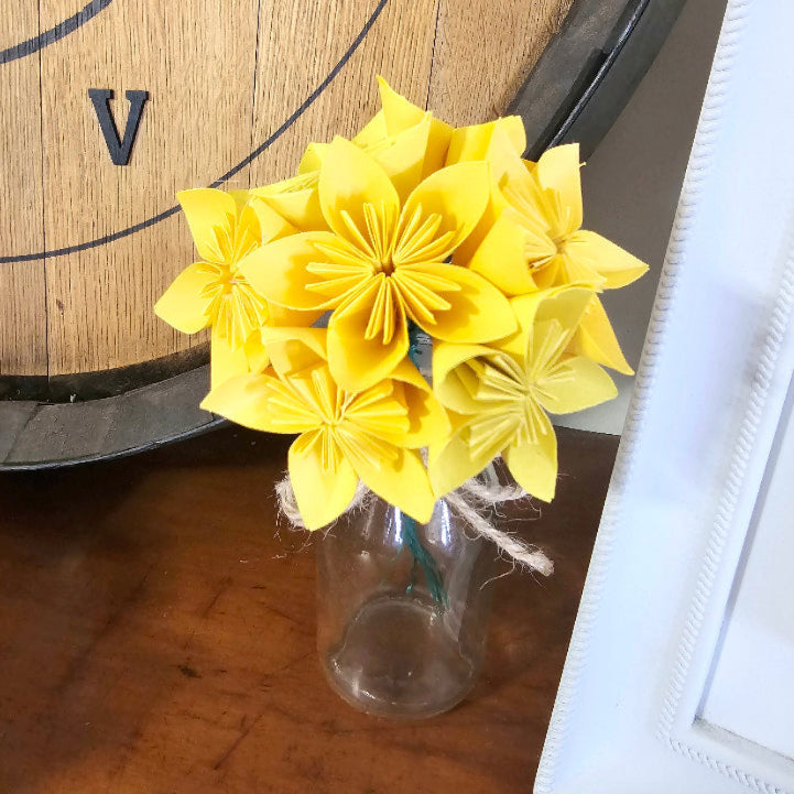 Yellow flowers in a clear glass bottle on a wooden surface with a clock in the background.